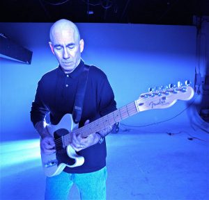 Jim standing playing guitar, blue lighted background.
