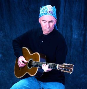 Jim sitting with blue background, playing acoustic guitar.